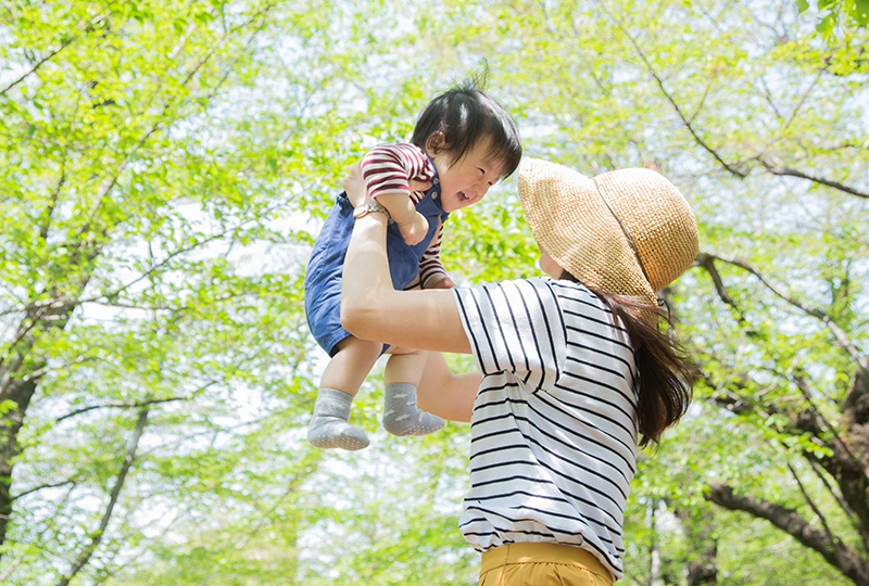 自然の中でママに高く持ち上げられ笑顔をみせる幼児の写真
