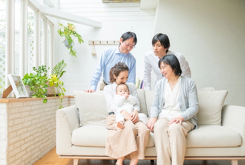親子3世代が母親に抱っこされる赤ちゃんを微笑ましく見る写真